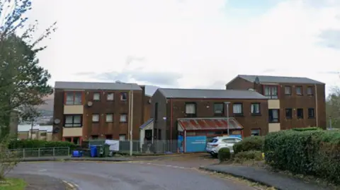 A picture of Kennedy Road in Fort William. The road curves around the left and behind it there are three brown buildings.