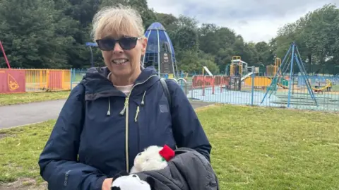 BBC/Claire Hamilton A blonde woman wearing sunglasses and carrying her granddaughter's white soft toy stands in front of the closed playground. 