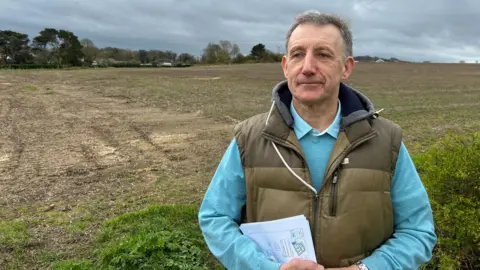 A man standing on the edge of a field, where there are plans to build 125 houses. He has receding grey hair, is looking to the left of the camera, and is holding a sheaf of papers which show the housing plans. He is wearing a khaki bodywarmer and a blue jumper.