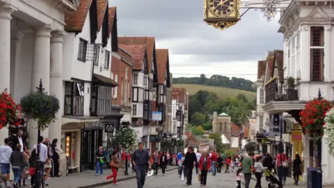 Guildford High Street, close to Tunsgate Arch. There are lots of shoppers on the street. Baskets of flowers are on either side of the road. The Guild Hall clock can be seen at the top of the picture and countryside in the background.