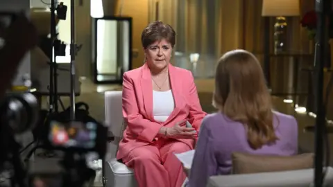 Nicola Sturgeon with her head tilted and a pensive look on her face. She is wearing a pink jacket and has a chain around her neck.