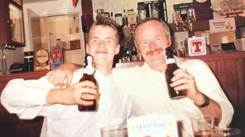 Shane Gorman Shane and David Gorman, wearing similar white shirts, their arms around one another, toast the camera with bottles of beer in a bar.