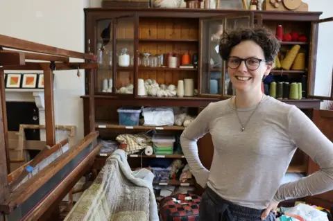 Emily McIlwaine smiling in a grey tshirt beside a large wooden loom with textiles across it
Lots of yarn and threads pictured on shelves in the background