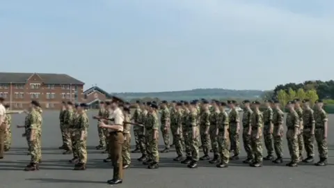 Family Handout A screenshot from a video showing a regiment practicing their uniform marching and saluting on camp at Larkhill. There is a large group of around 30 people dressed in green and brown camouflage uniforms and black caps. They are standing on a large tarmac area surrounded by fields and trees. In the background there is a brown brick building with lots of windows.