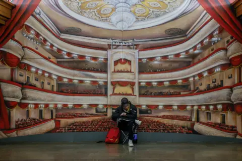 Alina Smutko/Reuters A person sits cross-legged in a metro station in Kyiv, in front of a mural depicting a large theatre, during an air raid in Kyiv in March 2024.