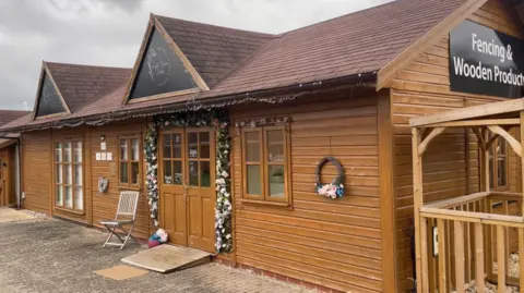 A wooden cabin-style building with a dark tiled roof houses the nail salon, with its entrance framed by an arch of pink and green artificial flowers. A wicker wreath hangs on the front wall and a small folding chair sits outside the door. A sign for fencing and wooden products is visible on an adjoining building to the right.
