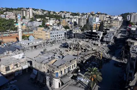 Reuters Destroyed market in Nabatieh. Gray debris from a pulverized structure covered a central square, which was surrounded by buildings with blown out windows. High-rise buildings stand on the hills in the background. 