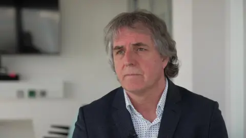 Steve Rotheram, who has grey hair and wears a dark suit, speaks to the camera