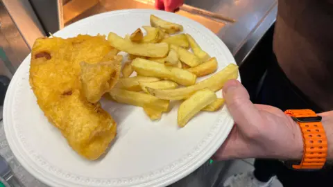 John Devine/BBC A white dinner plate with fish and chips on it, a fryer is visible too, in a fish and chip shop.