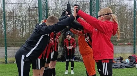 Angela Heap / Rothwell Juniors FC Members of a junior football club wearing red and black coloured football kit. The young football players have formed a human tunnel by joining their hands together and one of their team mates is stood at the entrance of the tunnel.