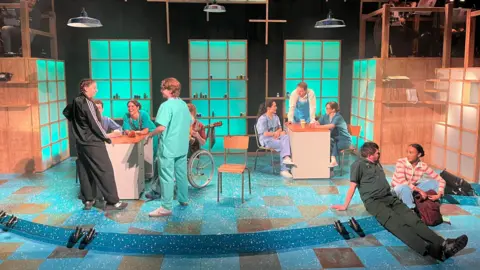 A wide shot of the Lifeline stage set, featuring wooden grid-like structures with teal glowing panels. Multiple performers are dressed in medical scrubs and professional clothing, interacting across the stage.