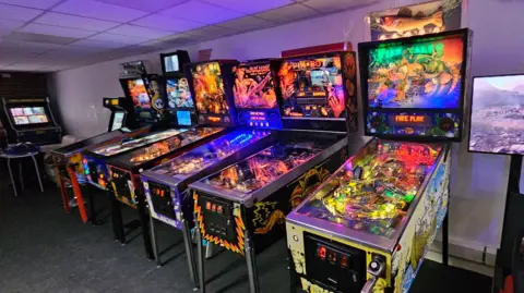 Six retro pinball machines in a row in a room, all very brightly coloured and lit up with internal lights.