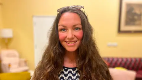 Rebekah Hester smiling at the camera. She has long brown hair and sunglasses perched on the top of her head, and is stood in a room with yellow walls.