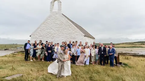 This image shows a couple after getting married, they are stood outside the little church with their wedding guests stood behind them. 