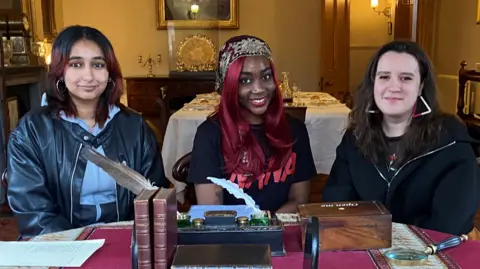 Elizabeth Gaskell's House Guruleen, Princess and Georgia smile as they site behind a desk with antique writing materials and books. They are in the Victorain dining room of Gaskell's House with table and dinner service items behind them.