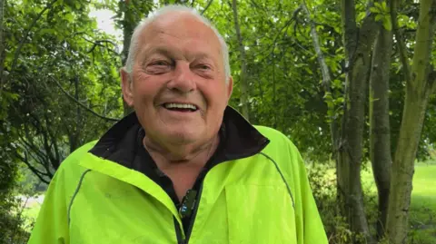 An older man with cropped receding white hair smiles at the camera. He's wearing a fluorescent yellow jacket, standing in front of trees.