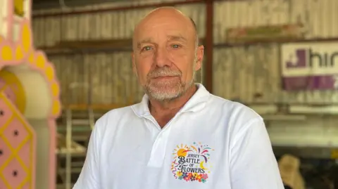 Martyn Maguire wearing a white Jersey Battle of Flowers polo shirt stands inside a workshop beside a large pink and white parade float structure. The workshop has metal walls and various equipment in the background.