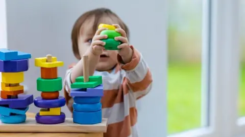 Getty Images A child playing with a wooden stacking toy with a window in the background. The child wears an orange and cream striped top and holds some of the wooden shapes in the air. 