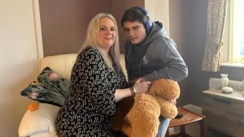 Charlotte Charlotte and Tobie pictured sitting in a living room. They share a large arm chair. She has a black and white dress on. He is wearing a grey hoodie and holding a large teddy bear. He is wearing blue ear protectors. 