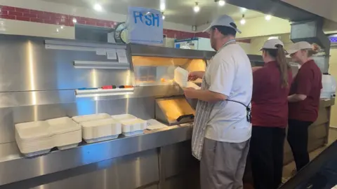 A man in white overalls and two women in burgundy polo shirts stood at a silver fish and chip fryer.