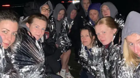 Andrew Harper A group of women huddle together at the side of the road in silver foil blankets