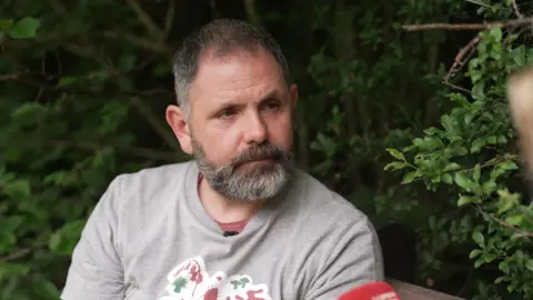 A man in a grey t-shirt looks seriously to the right of the camera. He is surrounded by green leaves.
