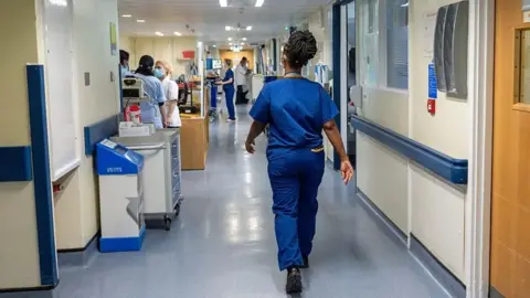 A woman wearing all-blue hospital scrubs walks away from us down a hospital corridor. On the floor is blue lining with other people in the distance.