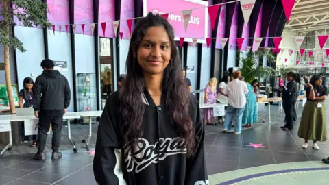 Emily Holt / BBC Amandeep stands in a college atrium wearing a black varsity-style jacket, with registration tables, pink bunting, and groups of people in the background