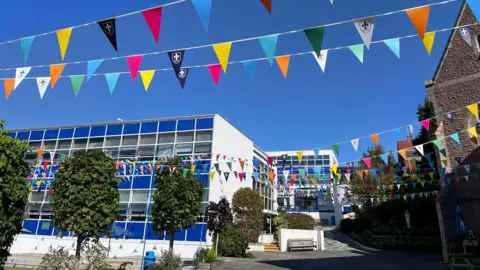 Beaulieu Convent School Beaulieu Convent School on a bright blue sky day. There are two buildings which are electric blue and feature many windows. There is multi-coloured bunting strung up high above trees and a bench.
