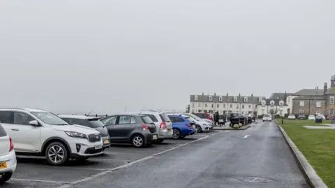 LDRS View down a road with a grassed area on one side and a line of cars parked in bays on the other.  The road ends at a junction, which is backed by a line of terraced houses. The ground is wet and it looks as if it has just stopped raining.