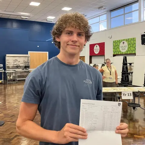A young bioy with curly brown hair smiling as he holds a sheet of paper in front of him.