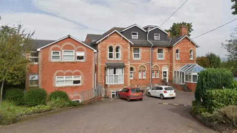 A large brick building surrounded by trees, with two cars parked outside. There is a small conservatory near the entrance door.