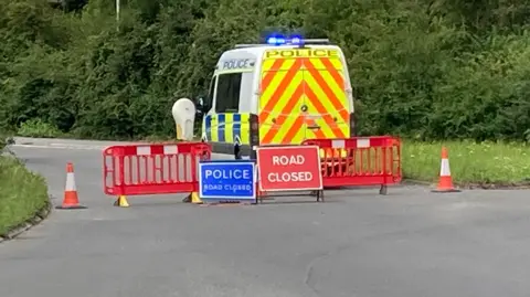 A police van parked in the middle of the road with its flashing lights on, along with red barriers, road closed signs and orange cones.