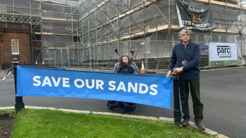 Two protesters against the Whitesands flooding scheme with a sign saying Save Our Sands. One woman is in a wheelchair and a mand with grey hair and glasses is standing beside the sign
