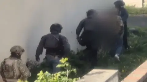 Blurred still from footage showing the backs of unidentifiable counter-terror officers arresting a man whose face cannot be seen. He is bent downwards walking along with the officers