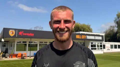 BBC/Jasmine Lowe A man smiling at the camera while standing on a green grass pitch under a blue sky. He has cropped fair hair, a matching beard and a tan. He is wearing a black Hull City training shirt and looks hot after a training session. A single-story building in the background has a sign reading "Hull City Training Ground."