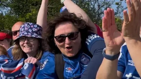 Connor Bennett/BBC Ipswich Town fans lean over a railing and celebrate the promotion. Many wear blue Ipswich Town football shirts and hold flags with the club's logo on them.