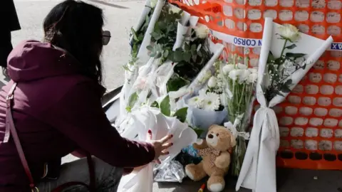 A woman seen in semi profile and wearing a maroon top and sunglasses lays a bouquet of flowers 