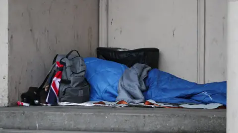 A blue tent depciting a rough sleeper with a grey rucksack nearby with part of a Union Jack flag showing attatched.