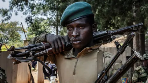 A member of the M23 movement in military fatigues carries weapons over his shoulder. Another fighter is to his right and he is holding weapons under his arm.