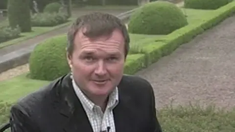 A younger Peter Henley, with brown hair flattened by rain, presents a TV programme from the ornate garden of Blenheim Palace. He wears a dark jacket and an open-necked checked shirt.