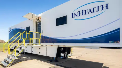 A mobile unit used to scan lung cancers - it is a large trailer with "In Health" written on the side and metal steps leading up to it with yellow handles.