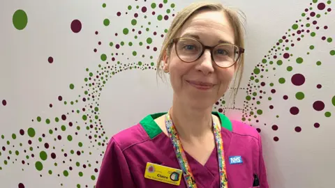 Ollie Conopo/BBC Woman with blonde hair and glasses smiling wearing a purple top with green around the neck. She also has a brightly-coloured lanyard and a badge saying Claire". She is standing in front of an artwork containing green and red dots.