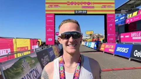 Sam Cook - a man in running sunglasses stands in front of the finish line. He has a white vest on and is wearing his medal.
