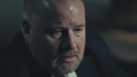 The Garden Productions A close up of Det Supt Iain Moor who has a greying close-cropped beard, is wearing a dark suit and is looking down and to the right. 