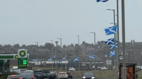 A row of blue and white saltire flags attached to lampposts on a busy road in Peterhead. Several cars are passing by.