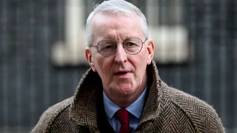 Northern Ireland Secretary Hilary Benn , he has short white hair, round-rimmed glasses and is wearing a brown herringbone-patterned overcoat over a blue shirt and red tie