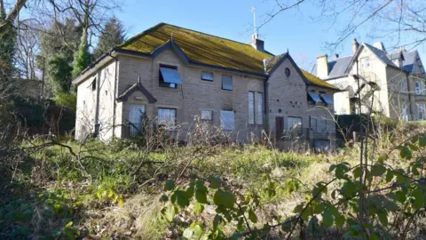 Image shows and abandoned two storey building in white stone with numerous windows with steel shutters over them. The building stands in an overgrown garden