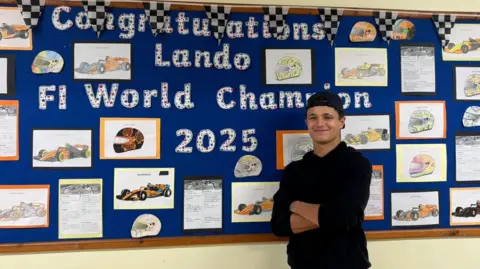 Lando Norris is standing in front of wall with drawings of his career by school children. The pictures have been stuck on by pins on to a blue board. Lando is a wearing a black reversed cap and a black jumper. 