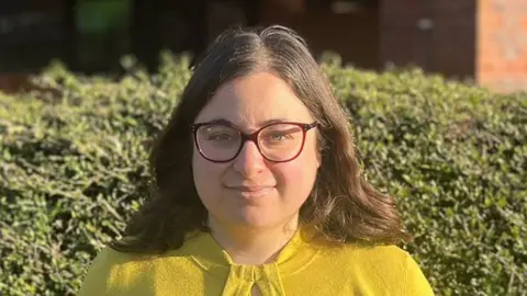 Emma Fielding Emma Fielding has shoulder-length brown hair. She is wearing a yellow top and glasses, and is standing in front of a bush, slightly smiling.